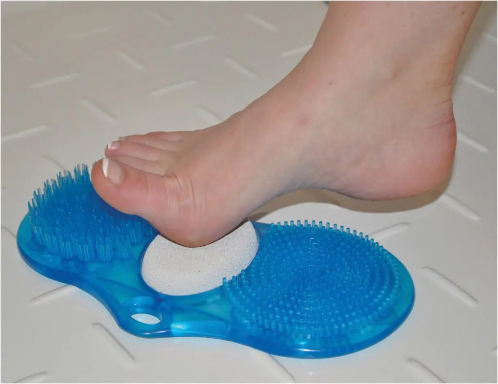 A bare foot rubs against the Foot Cleaner with pumice, a blue plastic scrubber with bristles and a pumice stone, on a white textured surface.