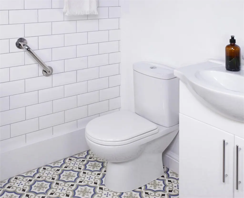 A white toilet beside a basin and cupboard, with a stainless steel grab rail in a polished finish mounted on the wall, and patterned floor tiles in a bathroom featuring white metro tile walls.