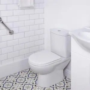 A white toilet beside a basin and cupboard, with a stainless steel grab rail in a polished finish mounted on the wall, and patterned floor tiles in a bathroom featuring white metro tile walls.