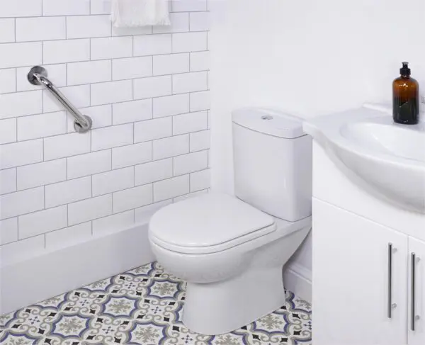 A white toilet beside a basin and cupboard, with a stainless steel grab rail in a polished finish mounted on the wall, and patterned floor tiles in a bathroom featuring white metro tile walls.