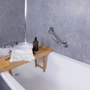 A white bath features a bamboo tray with a brown bottle and folded white towel, next to a wall-mounted stainless steel grab rail in a polished finish against grey tiled walls.
