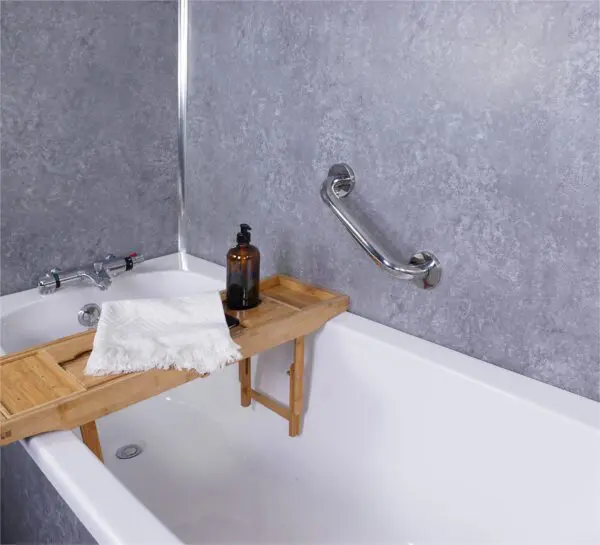 A white bath features a bamboo tray with a brown bottle and folded white towel, next to a wall-mounted stainless steel grab rail in a polished finish against grey tiled walls.