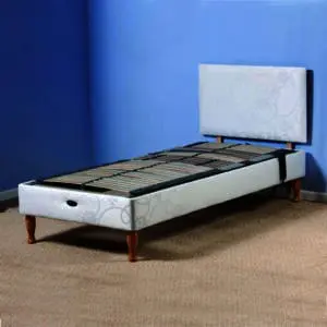 The Devon Electric Adjustable Bed features a white upholstered headboard and base, wooden legs, and exposed metal slats, shown on a beige carpet in a blue-walled room.