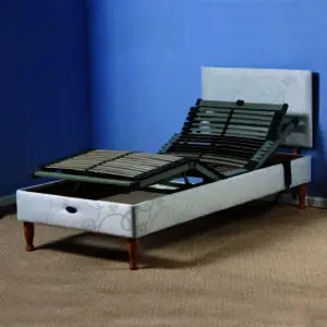 The Devon Electric Adjustable Bed, featuring a white frame and wooden legs, is shown with raised head and leg sections in a carpeted room with blue walls.