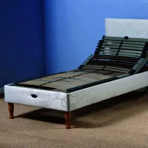 The Devon Electric Adjustable Bed features a raised head section, wooden legs, and a white upholstered frame, positioned on a carpeted floor against a blue wall.