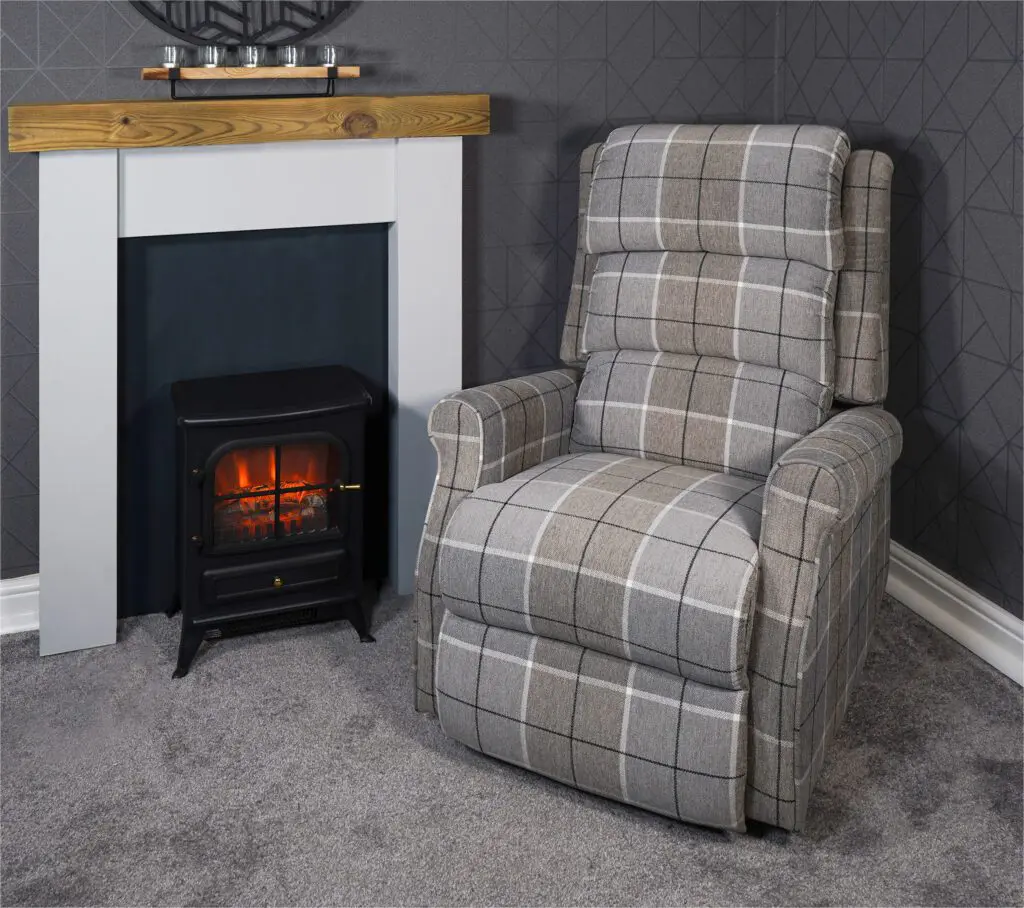 The Broadway Dual Motor Rise and Recline Chair sits next to an electric fireplace in a carpeted room with grey patterned walls and a wooden mantelpiece.