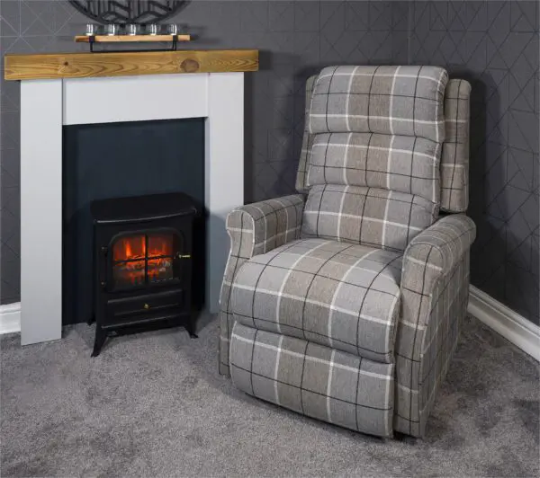 The Broadway Dual Motor Rise and Recline Chair sits next to an electric fireplace in a carpeted room with grey patterned walls and a wooden mantelpiece.