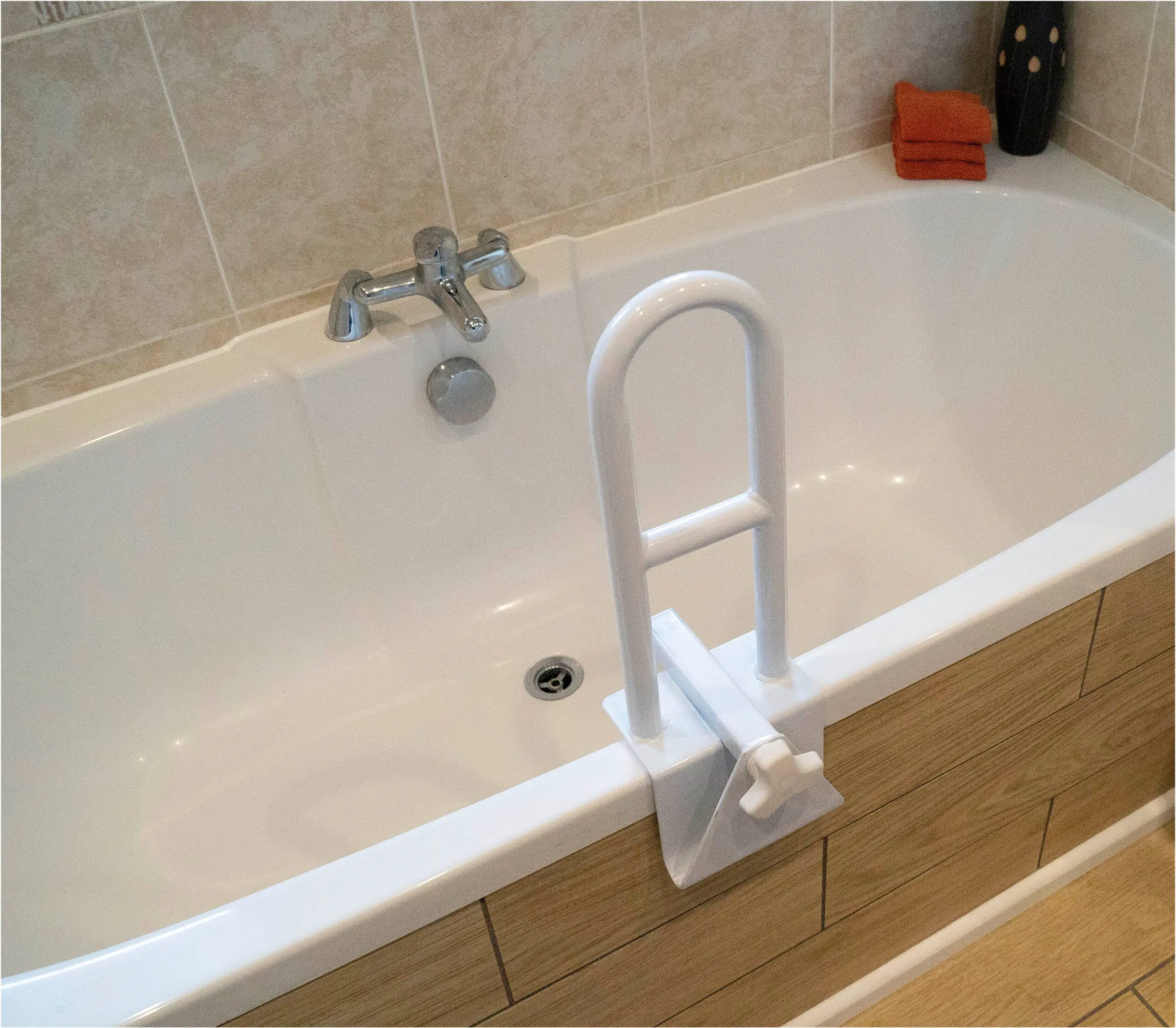 A white bath safety rail is attached to the side of a bath with a chrome tap. Orange folded towels and a decorative item are on the rim at the far end.