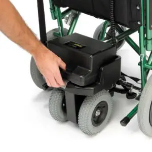 A hand is removing a black battery pack from the back of a green and black S Drive Powerstroll wheelchair with grey tyres.