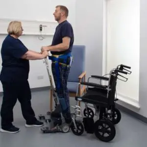 A healthcare worker assists a man using the Motion Transfer Aid next to a wheelchair in a clinical setting.