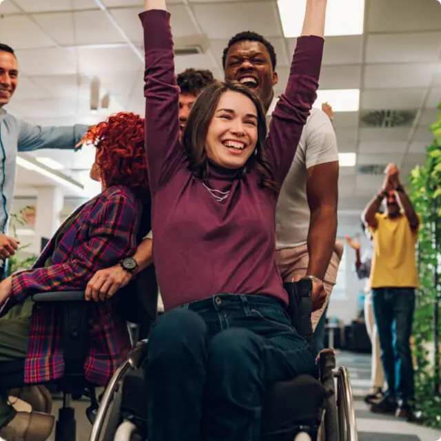 A woman in a wheelchair smiles and raises her arms in celebration, surrounded by cheering colleagues in an office setting that embraces Independent Living.