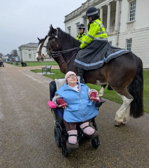 A woman in a wheelchair smiles during a Day Support outing as two police officers on horseback stand nearby in front of a large white building.