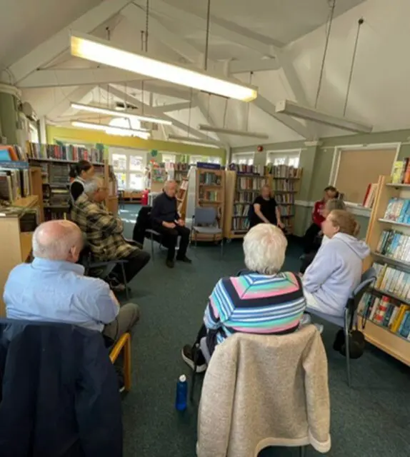 A group of people sit in a circle on chairs in a library, engaged in a Day Support discussion. Bookcases line the walls, and natural light streams through the windows.