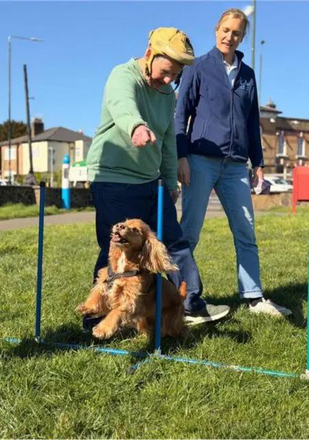 A man wearing a helmet, part of a Day Support programme, guides a small brown dog through an agility obstacle, while a woman stands nearby watching on a sunny day.