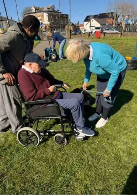 An older man in a wheelchair enjoys the outdoors on a grassy area, accompanied by two people as part of Day Support, with buildings and others visible in the background.