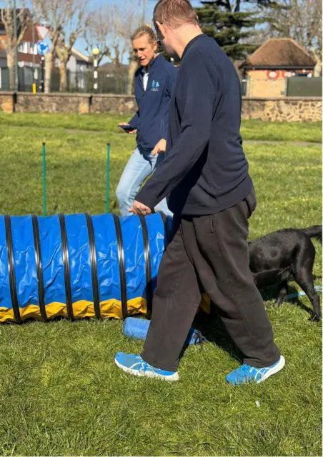 A man and woman with a black dog near a blue and yellow agility tunnel on a grassy field, enjoying Day Support activities, with trees and buildings in the background.