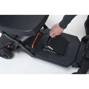 A person removes a rectangular battery from the base of a black mobility scooter with orange trim details.