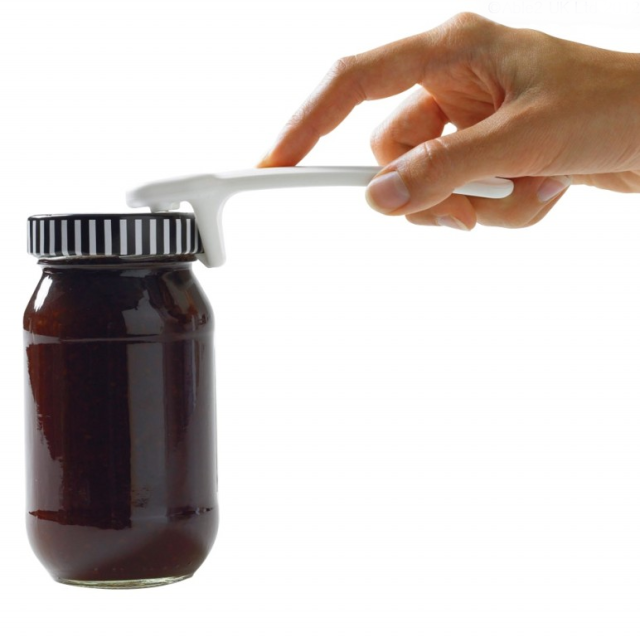 A hand uses a Jar Key to grip the lid of a sealed glass jar filled with a dark substance, against a white background.