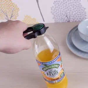 A hand uses a Plastic Multi Opener to twist the green cap off a Robinsons orange and pineapple juice bottle beside stacked blue dishes.