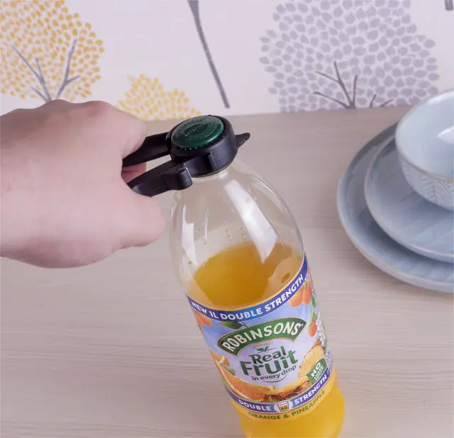 A hand uses a Plastic Multi Opener to twist the green cap off a Robinsons orange and pineapple juice bottle beside stacked blue dishes.