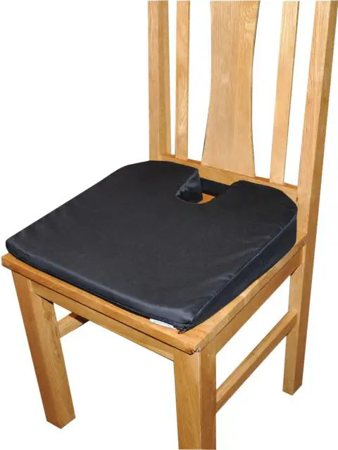 A wooden chair with a Coccyx Wedge Cushion featuring a black seat and U-shaped cut-out at the back.