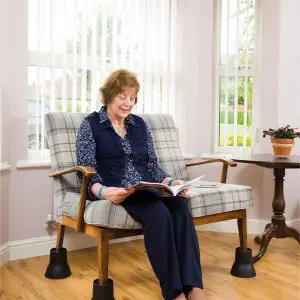 An older woman sits on a checked sofa elevated with Furniture Raisers (Set of 4), reading a magazine in a bright room with large windows, wooden floors, a side table, and a potted plant.