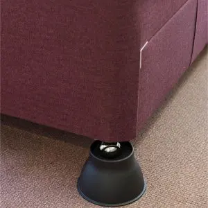 A bed with a maroon fabric frame is supported by a black plastic raiser placed under one of its legs, standing on a carpeted floor.
