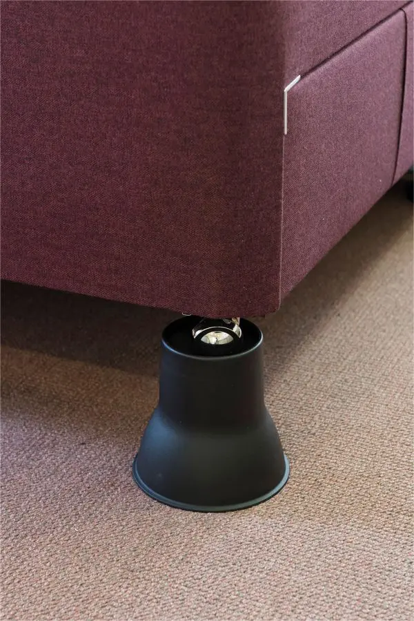 A close-up of a sofa leg placed inside a black plastic raiser on a carpeted floor.