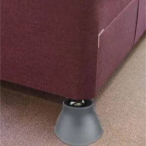 A close-up of a bed with a maroon upholstered frame, supported by a grey bed raiser placed under the leg, on a carpeted floor.