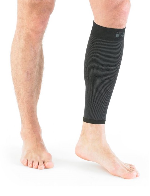 A person stands barefoot, wearing a black compression sleeve on their right calf, whilst their left leg is bare against a white background.