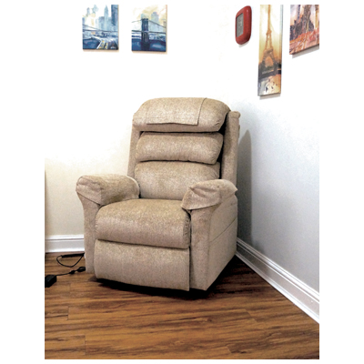 A beige recliner chair is positioned in the corner of a room with wooden flooring and framed art on the white walls.