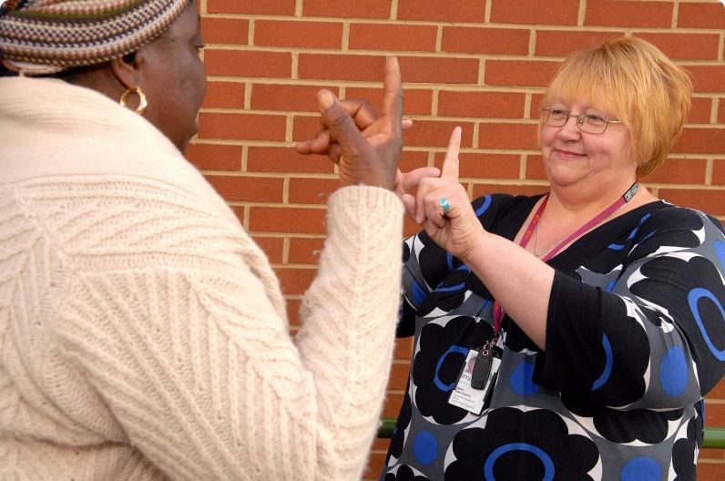 Two women are communicating using sign language in front of a brick wall; one is gesturing with her hands whilst the other looks on and responds.