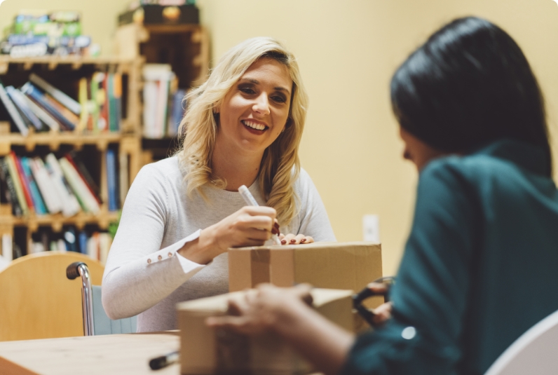 Two women sit at a table; one is smiling and writing on a cardboard box while the other faces away, also handling a box. Bookcases are in the background.