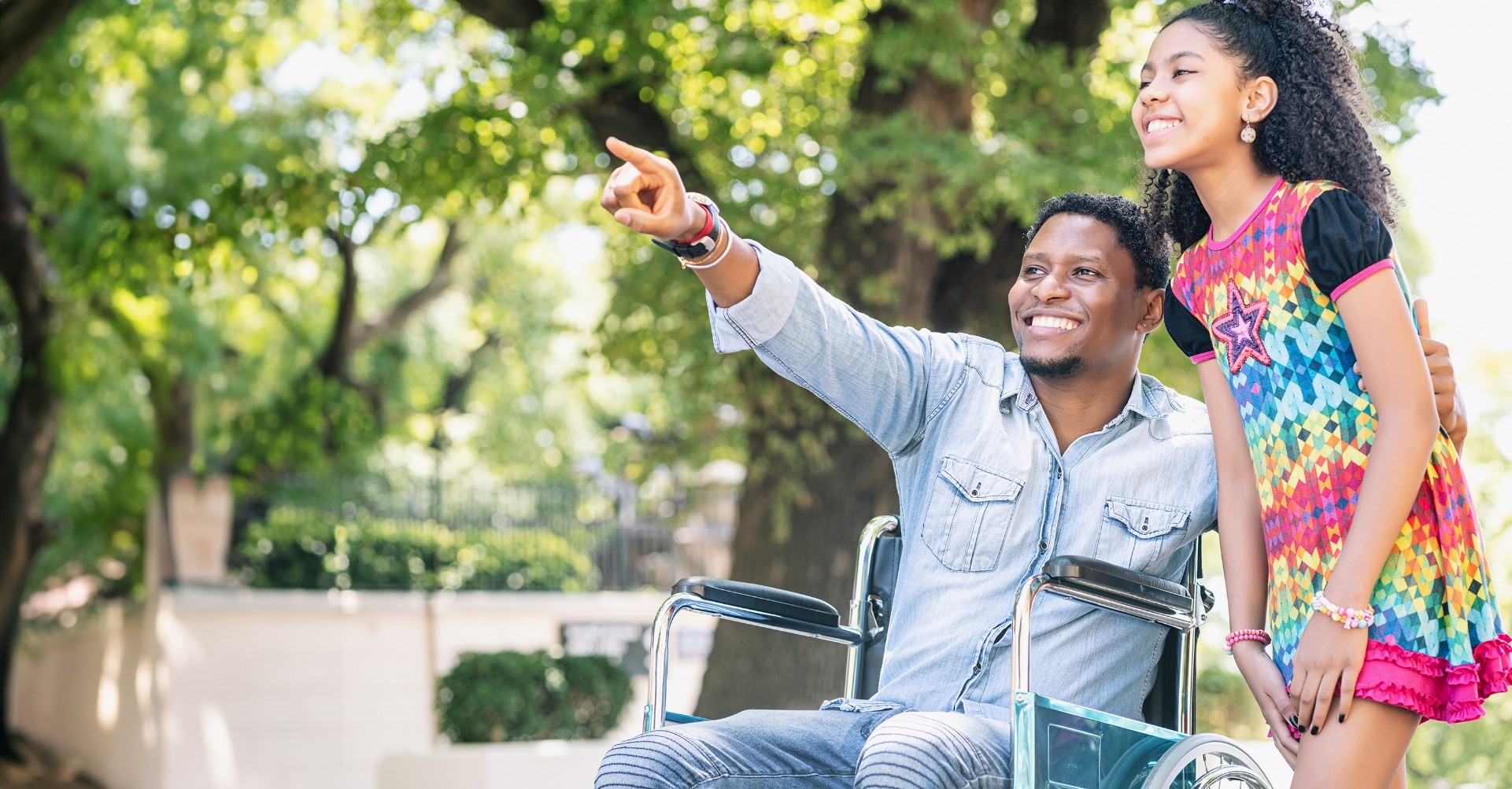 A man in a wheelchair points at something while smiling beside a young girl outdoors on a sunny day.