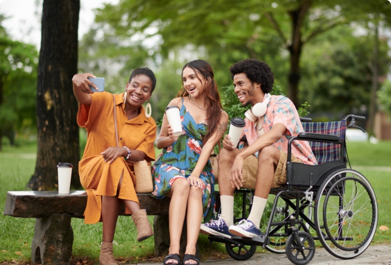 Three friends sit together outdoors; one takes a selfie, another holds a drink, and the third, wearing headphones, sits in a wheelchair. All are smiling.
