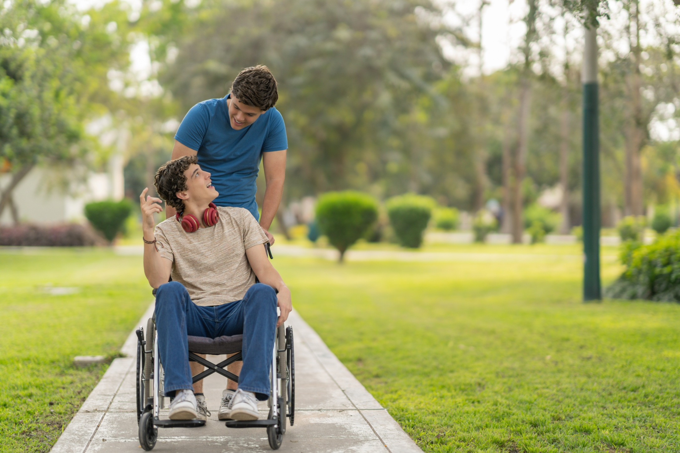 Two young men are outdoors; one is smiling and sitting in a wheelchair whilst the other stands behind him. Both appear to be enjoying a conversation on a park walkway, highlighting the joy of independent living.