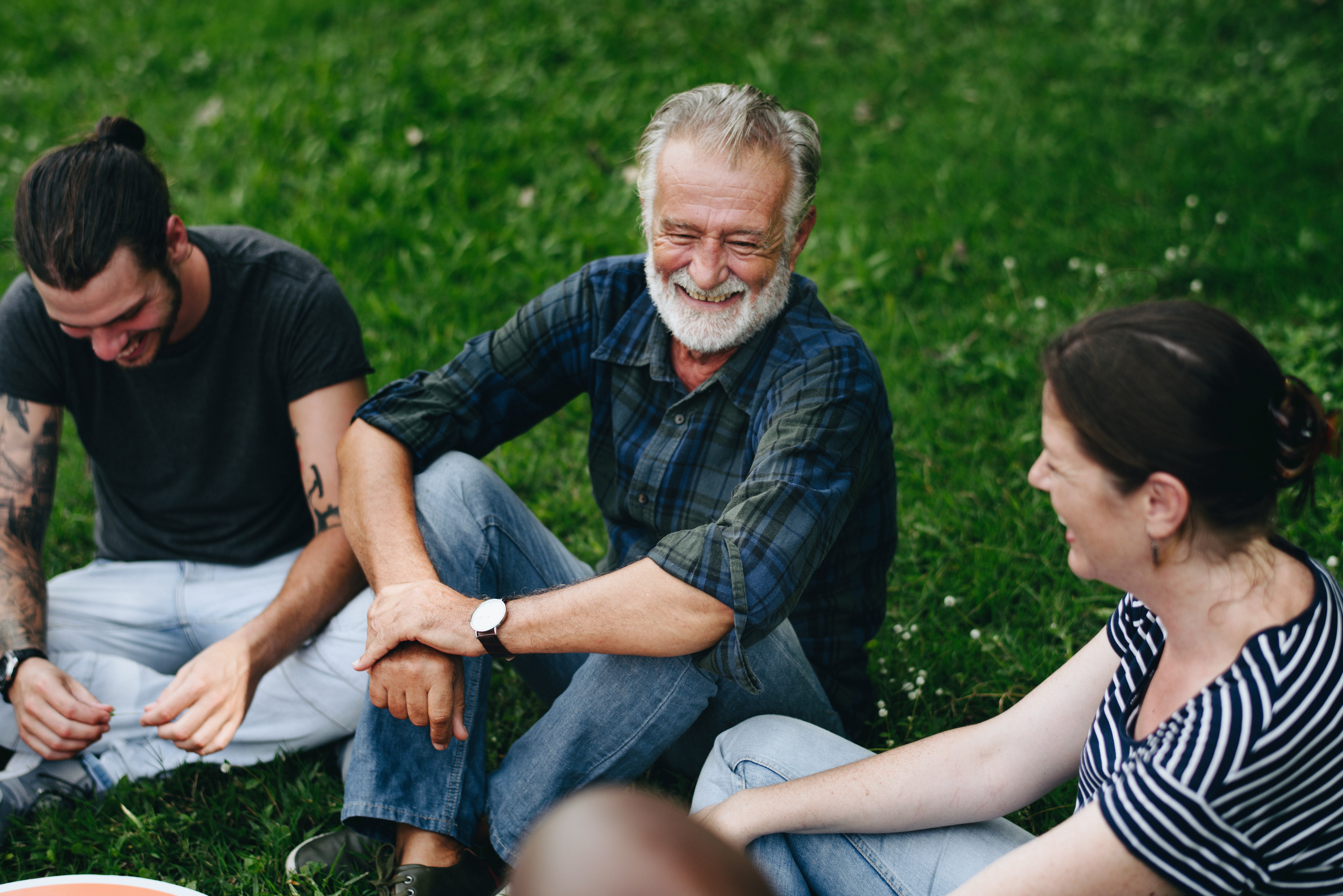Three adults sit together on grass, talking and smiling. The man in the centre has grey hair and a beard; the group appears relaxed and engaged in conversation outdoors.