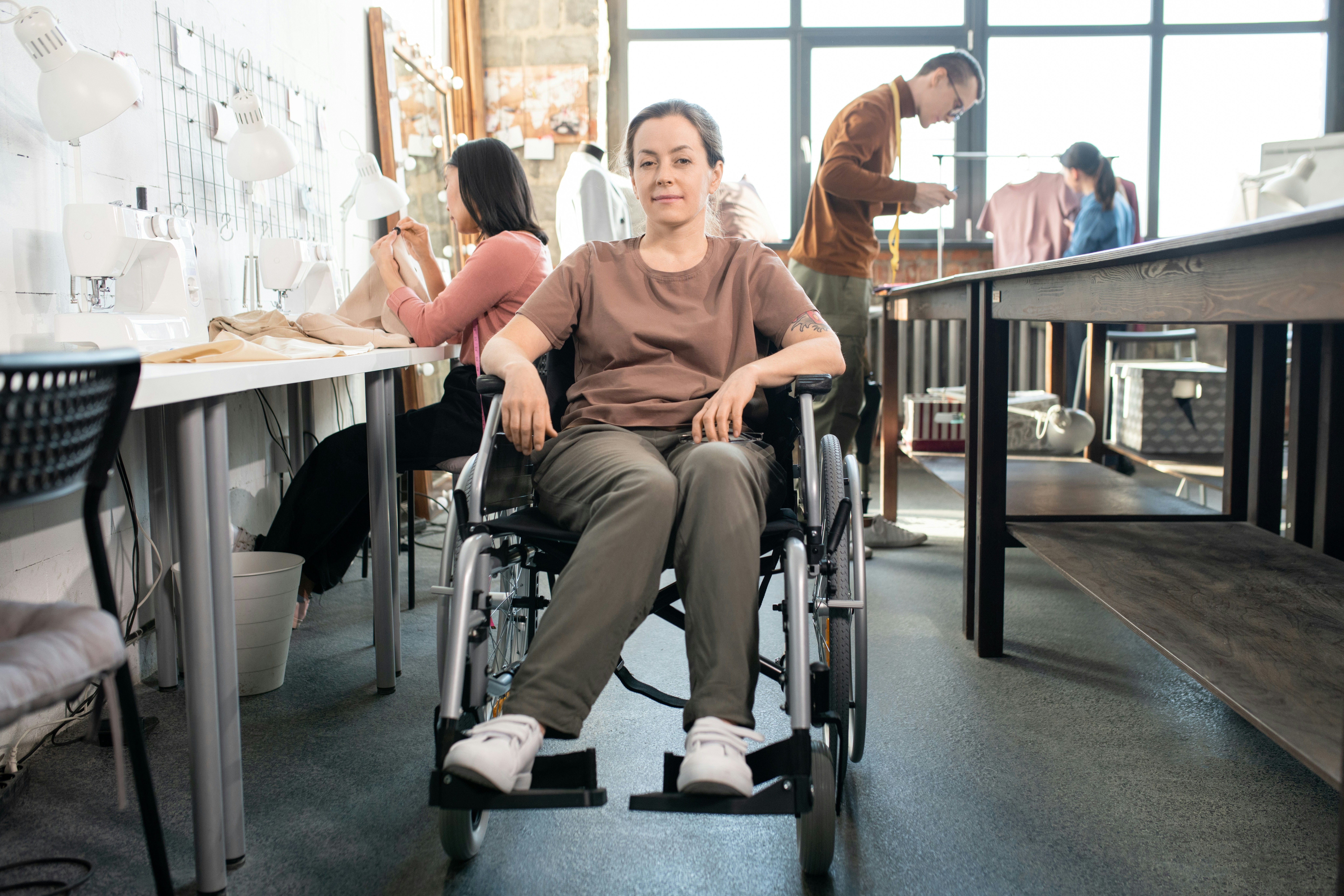 A woman in a wheelchair sits in a sewing workshop, with other people working at tables and sewing machines in the background.
