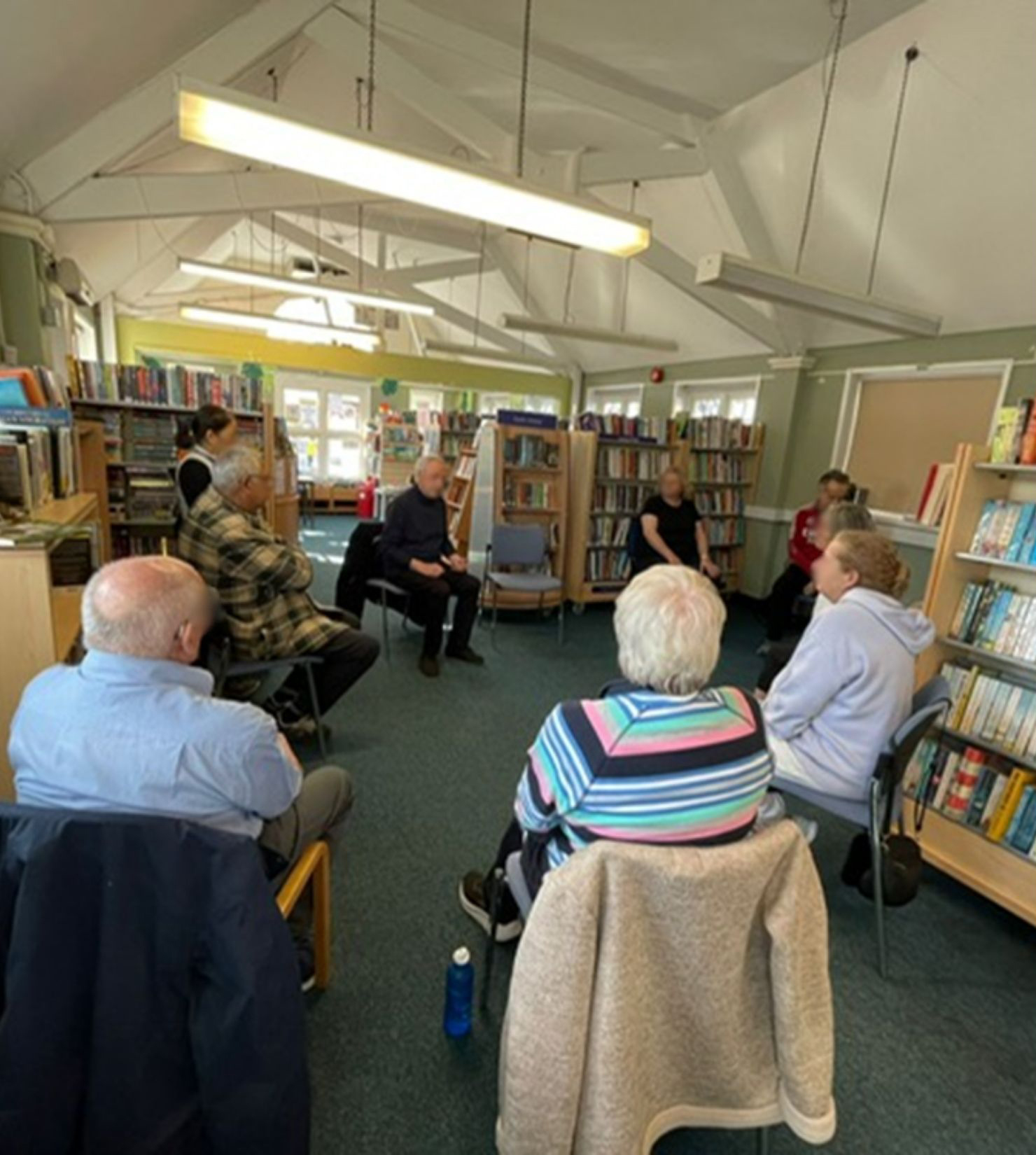 A group of people sit in a circle on chairs in a library, engaged in a Day Support discussion. Bookcases line the walls, and natural light streams through the windows.