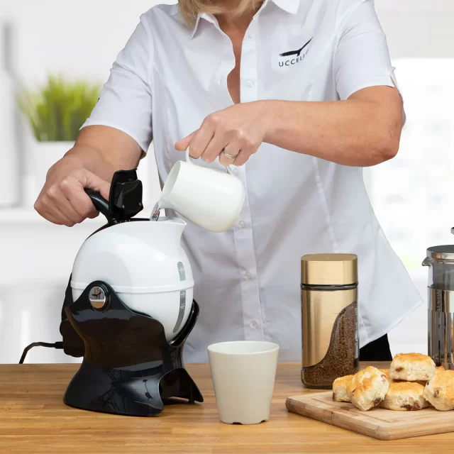 A person pours water from a jug into a Uccello Kettle on a kitchen worktop, with a cup, coffee jar, pastries, and a cafetière nearby.
