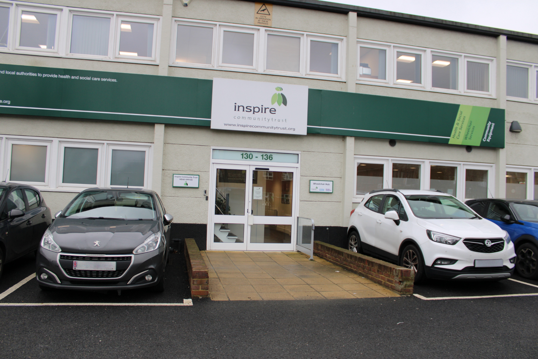Two cars parked outside the entrance of a building with a sign reading "Inspire Community Trust" above the door, where essential Community Equipment is provided to support local residents.