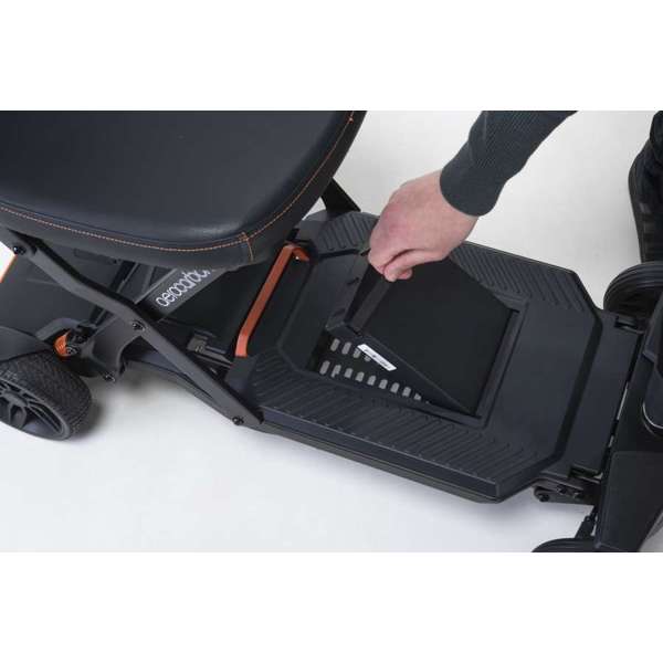 A person removes a rectangular battery from the base of a black mobility scooter with orange trim details.