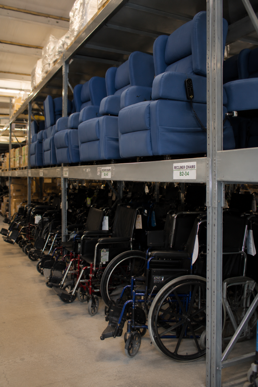 Multiple rows of blue recliner chairs are stacked on upper shelves, whilst various Community Equipment wheelchairs are lined up below in a warehouse storage area.