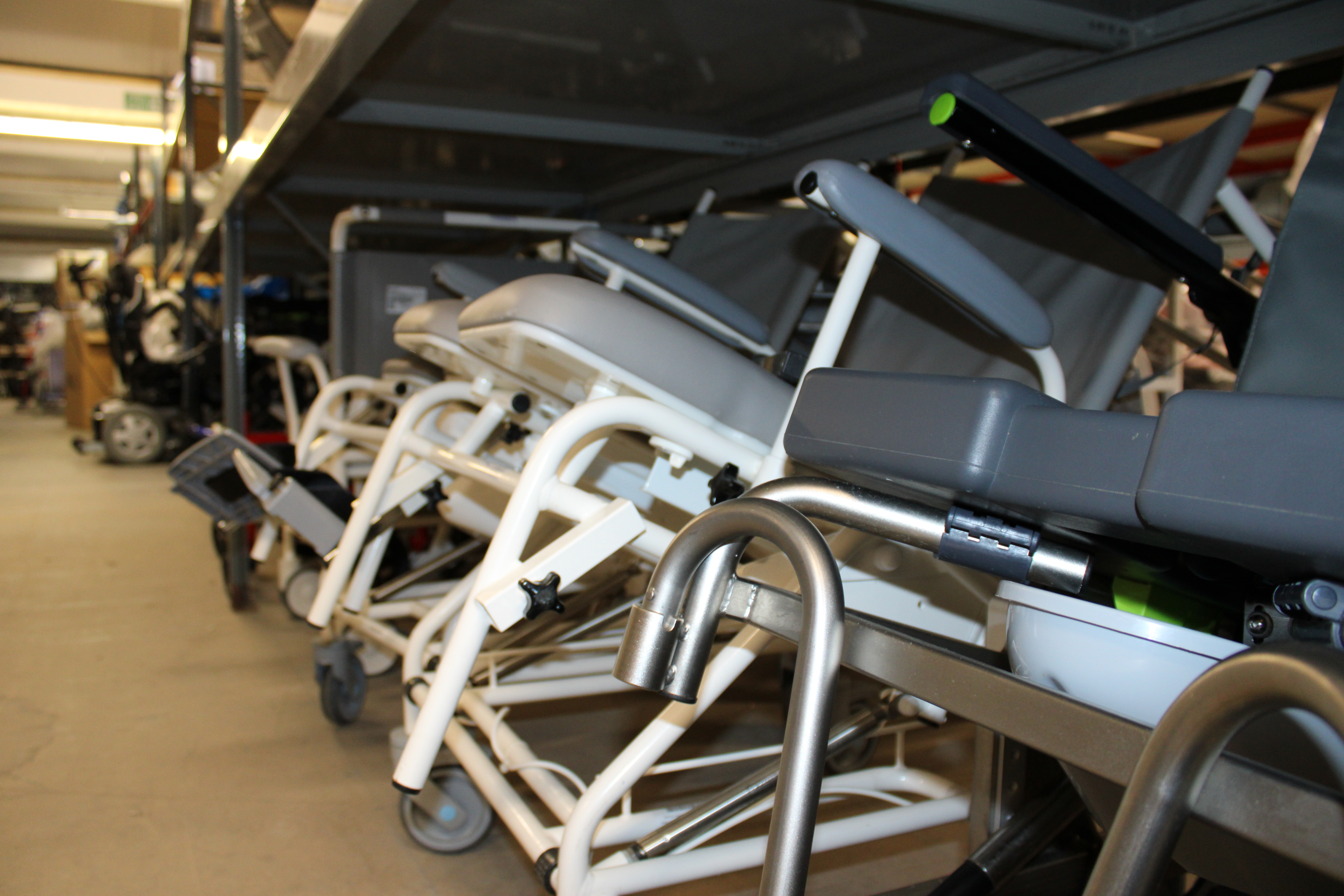 Several community equipment items, including wheelchairs and mobility aids, are stacked and stored on metal shelving in a warehouse or storage facility.