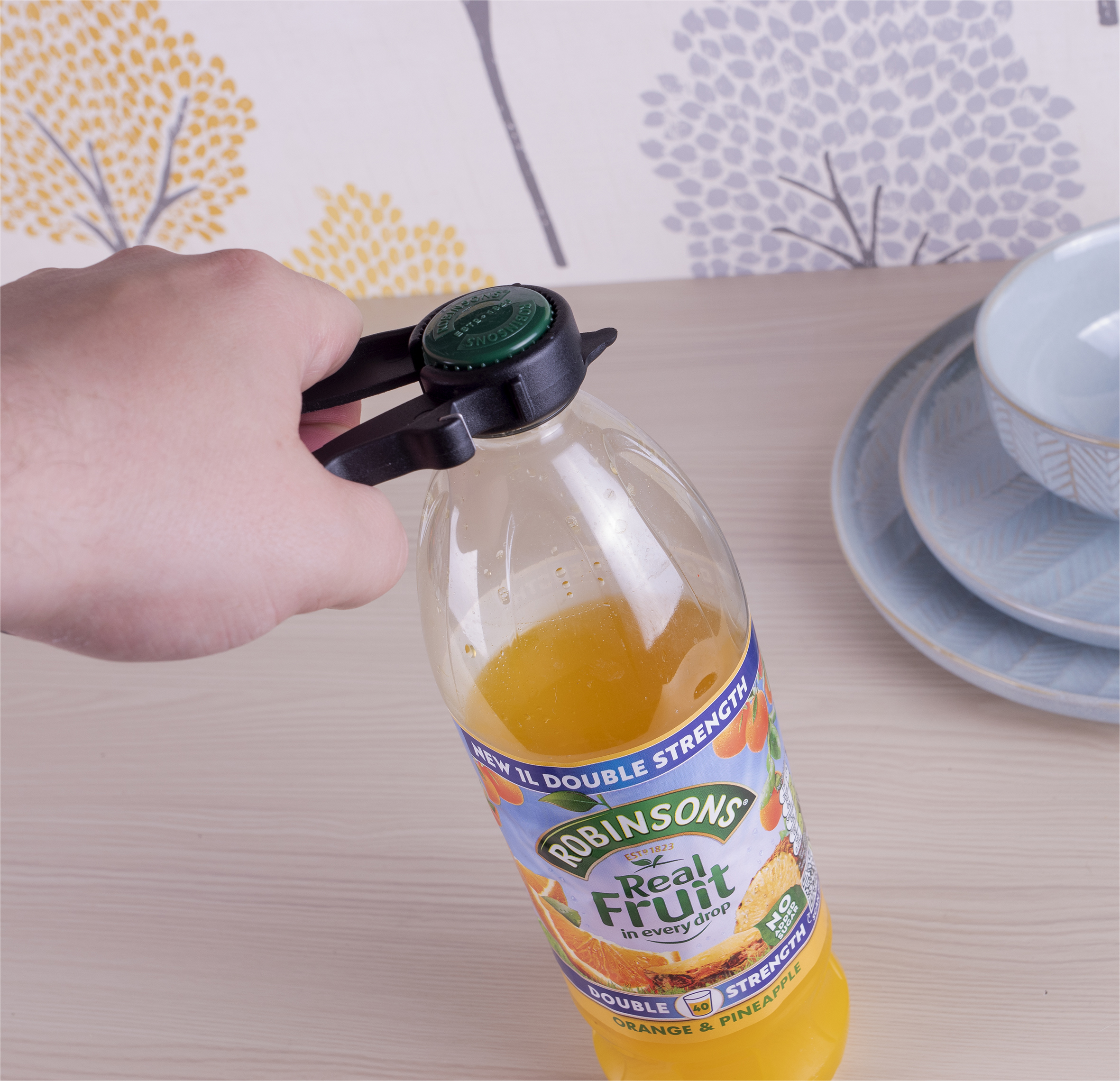 A hand uses a Plastic Multi Opener to twist the green cap off a Robinsons orange and pineapple juice bottle beside stacked blue dishes.