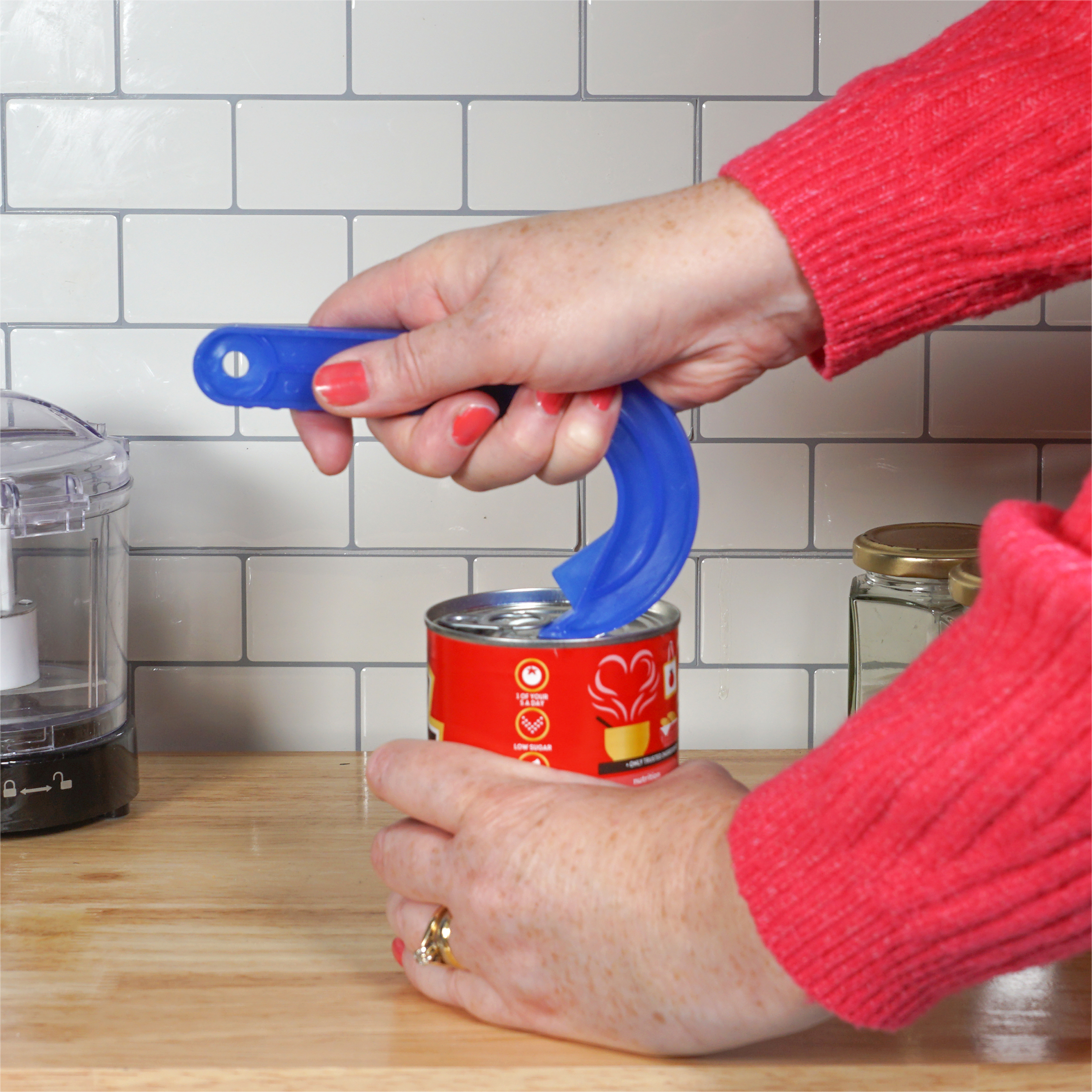 A person uses a Ring Pull Tin Opener to open a tinned food item on a wooden kitchen worktop in front of a white tiled splashback.