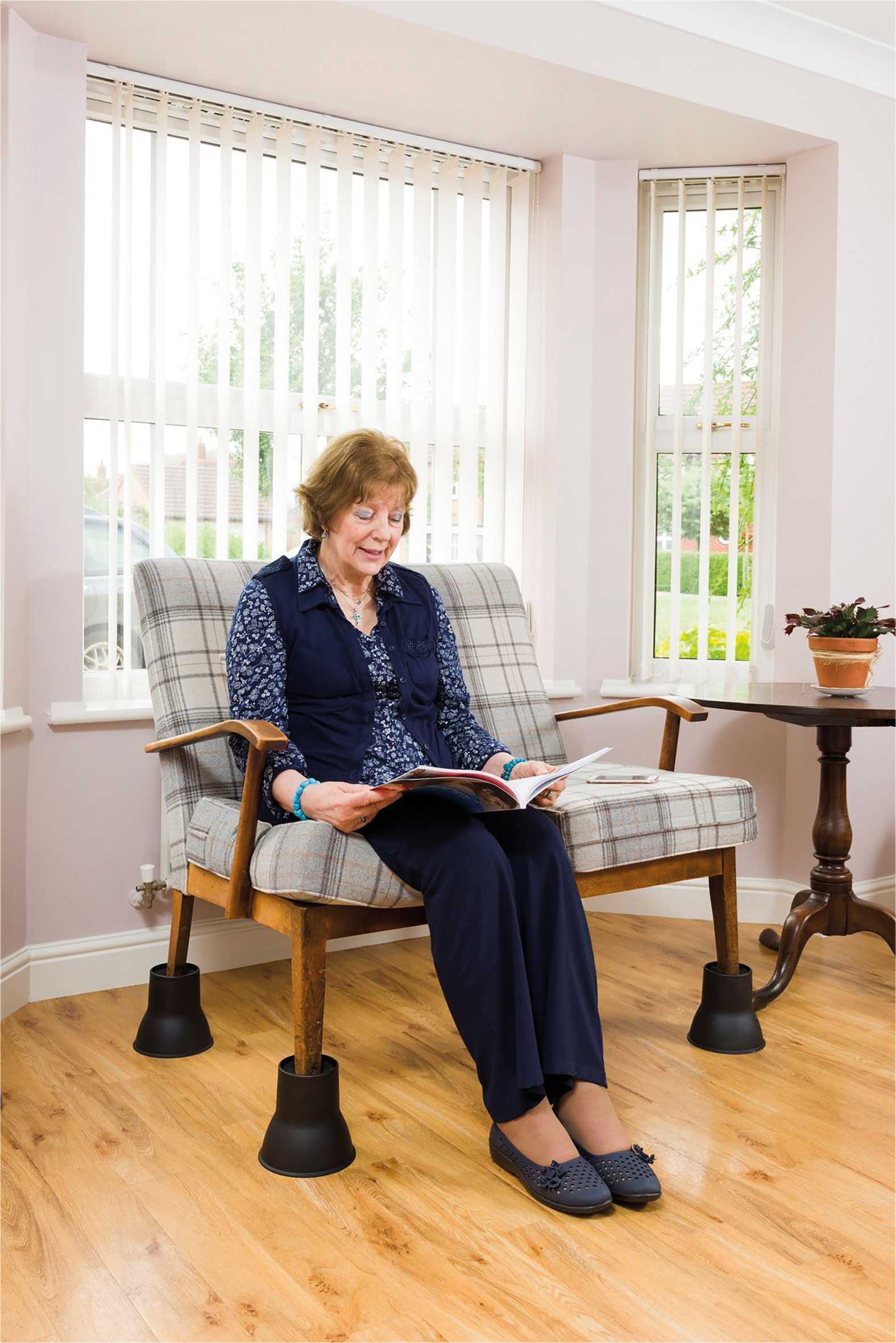 An older woman sits on a checked sofa elevated with Furniture Raisers (Set of 4), reading a magazine in a bright room with large windows, wooden floors, a side table, and a potted plant.