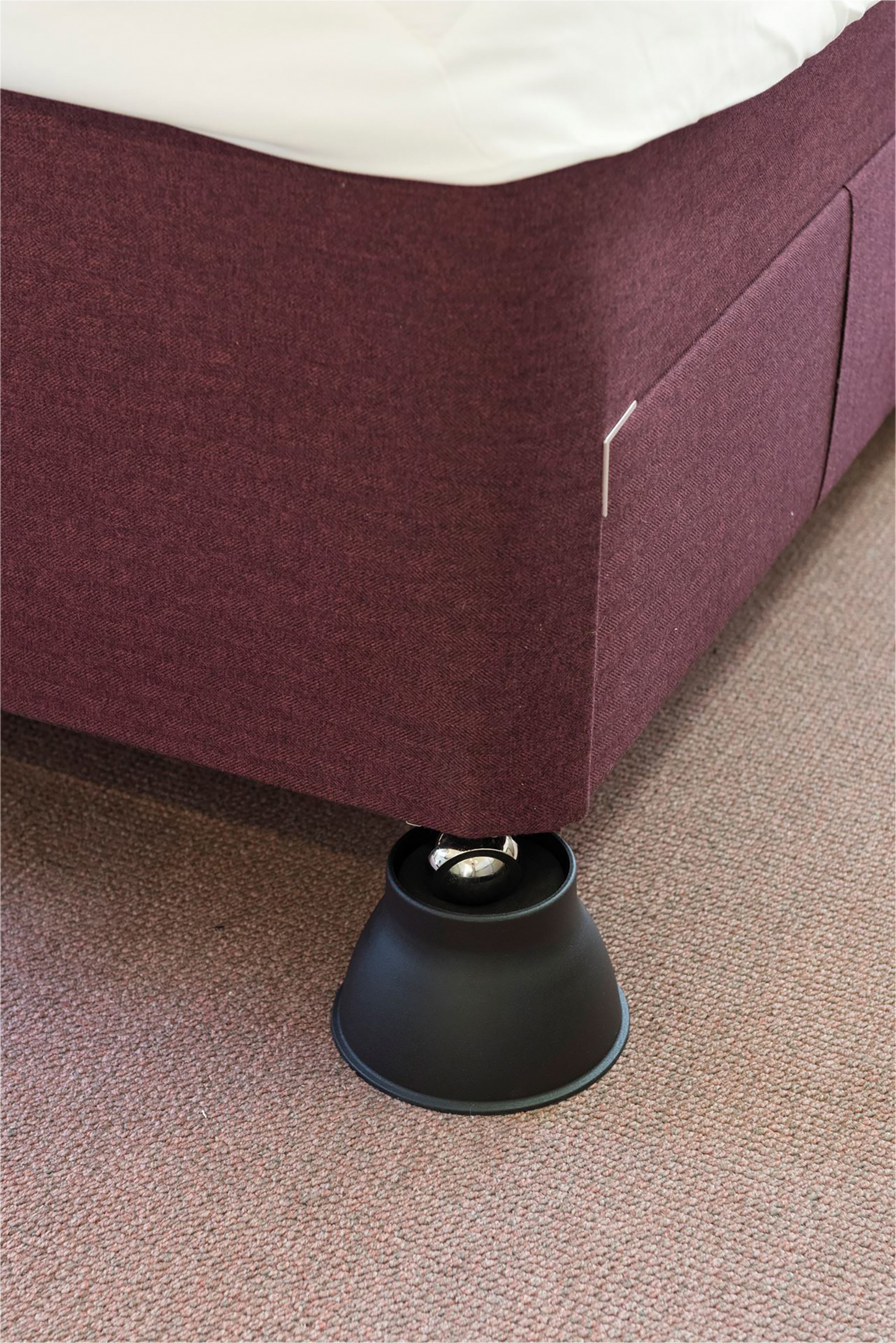 A bed with a maroon fabric frame is supported by a black plastic raiser placed under one of its legs, standing on a carpeted floor.