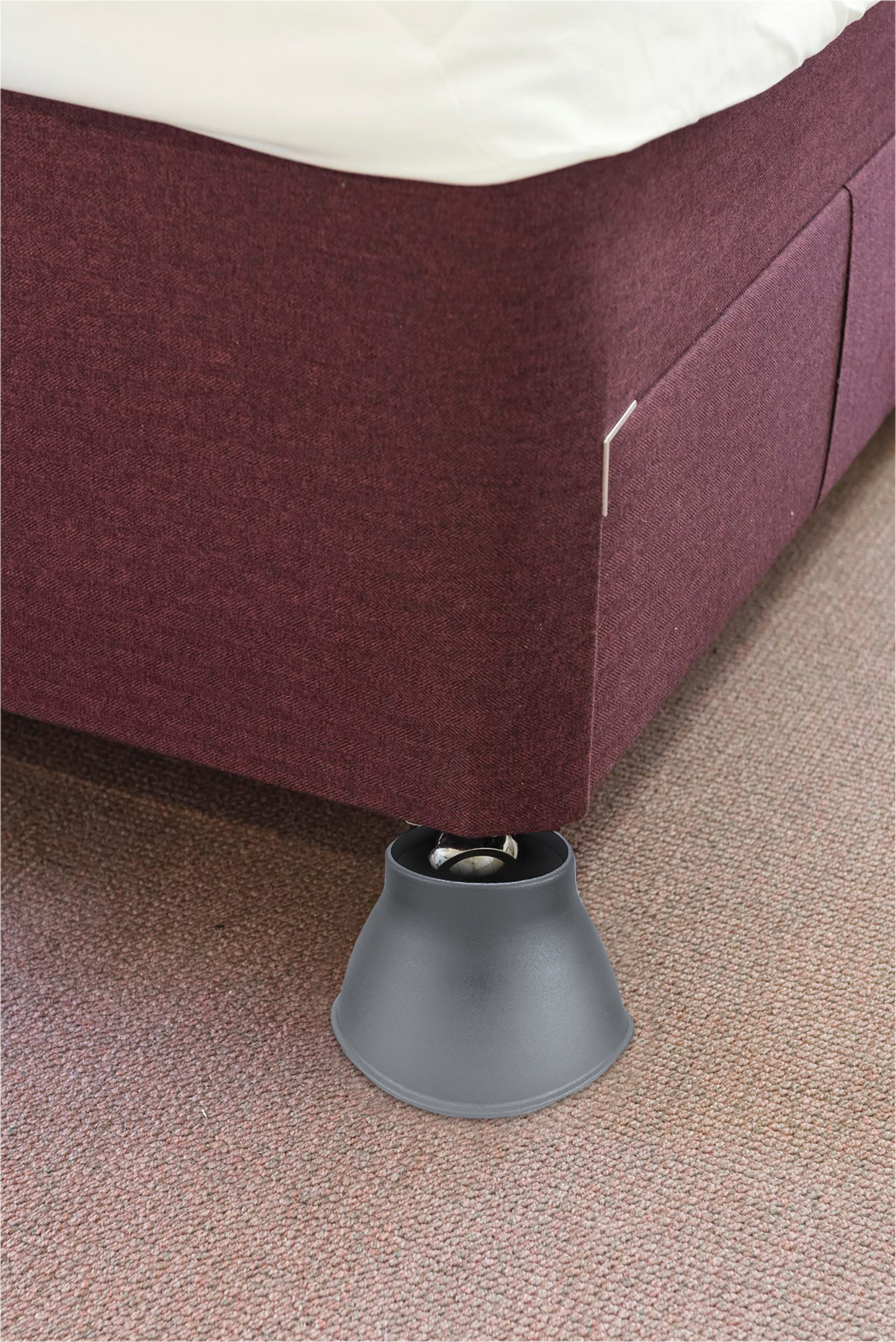 A close-up of a bed with a maroon upholstered frame, supported by a grey bed raiser placed under the leg, on a carpeted floor.
